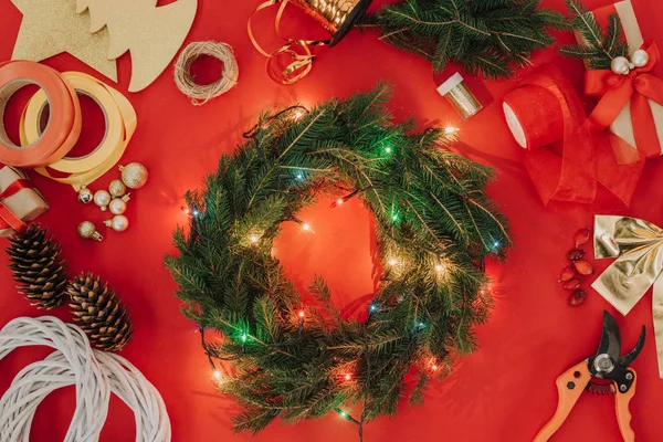 Flat lay with pine tree branches, christmas lights and ribbons for handmade christmas wreath on red backdrop — Stock Photo