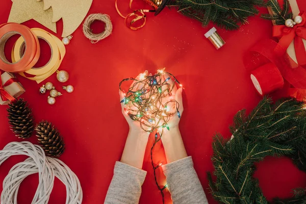 Schnappschuss einer Frau mit Weihnachtsbeleuchtung in der Hand, Tannenzweigen und Dekorationen für handgefertigten Kranz auf rotem Hintergrund — Stockfoto