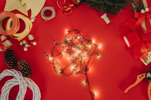 Flat lay com galhos de pinheiros, luzes de Natal e fitas para grinalda de Natal artesanal em pano de fundo vermelho — Fotografia de Stock