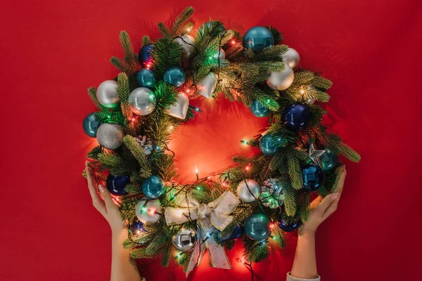 Partial view of woman holding handmade christmas wreath with lights and toys on red background — Stock Photo