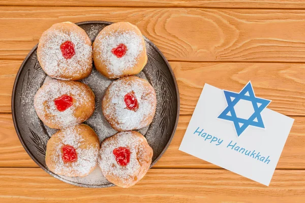 Vista superior de rosquillas dulces y feliz tarjeta hannukah en la mesa de madera, concepto de celebración hannukah - foto de stock