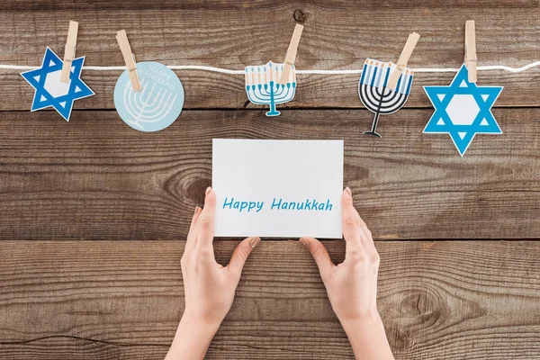 Cropped shot of woman holding happy hannukah card with holiday paper signs peged on rope on wooden surface, hannukah concept — Stock Photo