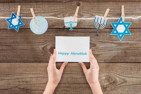 Recortado tiro de mujer sosteniendo feliz tarjeta hannukah con signos de papel de vacaciones pegado a la cuerda en la superficie de madera, concepto hannukah - foto de stock