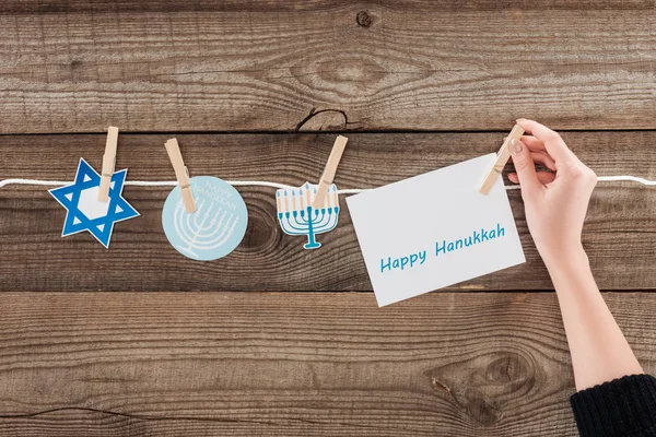 Recortado tiro de mujer vinculación feliz tarjeta hannukah en la cuerda con signos de papel de vacaciones en la superficie de madera, concepto hannukah - foto de stock
