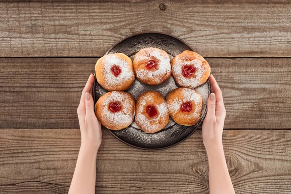 Vista parziale della donna che tiene piatto con ciambelle dolci hannukah cibo tradizionale su superficie di legno, concetto di hannukah — Foto stock