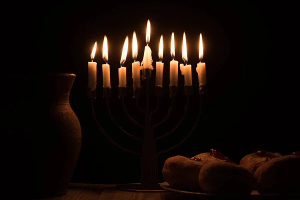 Close up view of traditional sweet doughnuts, clay jug and menorah with candles on black background, hannukah holiday concept — Stock Photo