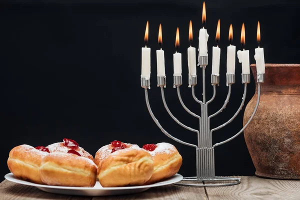 Close up view of traditional sweet doughnuts, clay jug and menorah with candles isolated on black, hannukah holiday concept — Stock Photo