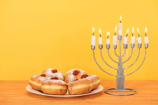 Close up view of sweet doughnuts and menorah with candles on wooden surface isolated on yellow, hannukah concept — Stock Photo