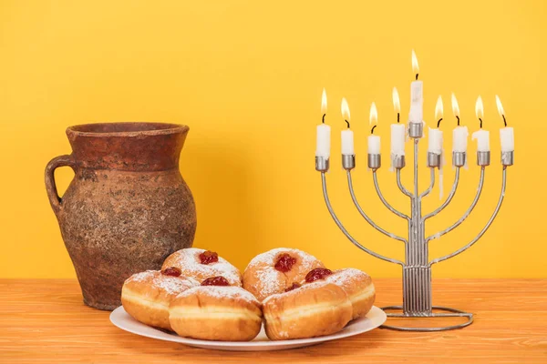 Nahaufnahme von süßen Donuts, Tonkrug und Menora auf hölzerner Oberfläche vor gelbem Hintergrund, hannukah-Konzept — Stockfoto