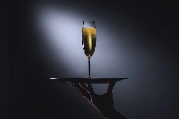 Vista parcial de la camarera sosteniendo bandeja de servir con copa de champán sobre fondo oscuro - foto de stock