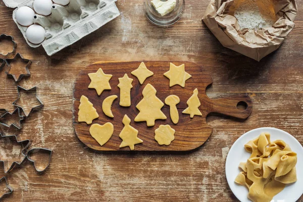 Flache Lage mit geschnittenem Rohteig zum Plätzchenbacken auf Schneidebrett auf Holzoberfläche — Stockfoto