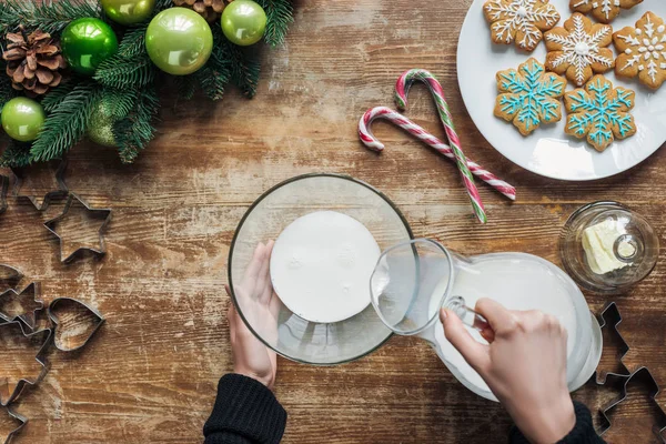 Tiro recortado de la mujer que vierte la leche en el tazón mientras que hace la masa para las galletas de Navidad en la superficie de madera - foto de stock