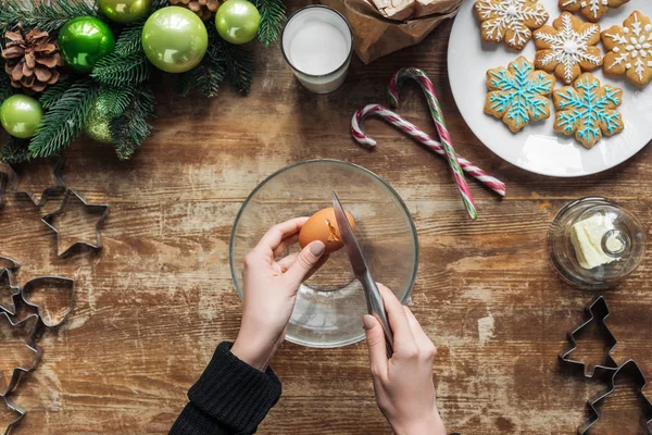 Abgeschnittene Aufnahme einer Frau, die Hühnerei in Schüssel knackt, während sie Teig für Weihnachtskekse auf Holztischplatte mit dekorativem Kranz backt — Stockfoto