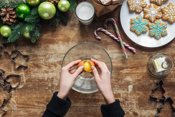 Abgeschnittene Aufnahme einer Frau, die Ei in Schüssel legt, während sie Teig für Weihnachtskekse auf hölzerner Tischplatte mit dekorativem Kranz backt — Stockfoto