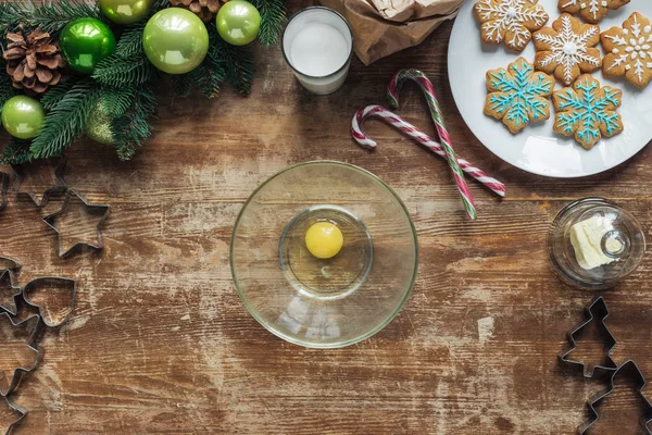 Vue du dessus de l'œuf de poulet cru dans un bol, biscuits de Noël sur une assiette et couronne de fête décorative sur une surface en bois — Photo de stock