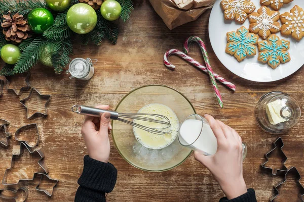 Abgeschnittene Aufnahme einer Frau, die Milch in eine Schüssel gießt, während sie Teig für Weihnachtsplätzchen auf einer hölzernen Tischplatte mit dekorativem Kranz backt — Stockfoto
