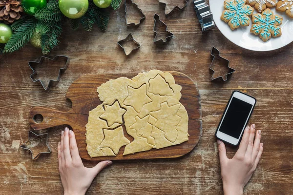 Vista parcial de la mujer, teléfono inteligente con pantalla en blanco y masa cruda para galletas de Navidad en la mesa de madera - foto de stock