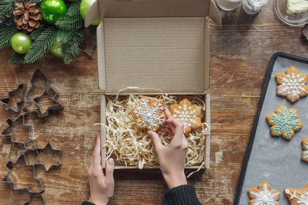 Обрезанный снимок женщины кладет домашнее рождественское печенье в коробку на деревянный стол с декоративным рождественским венком — стоковое фото