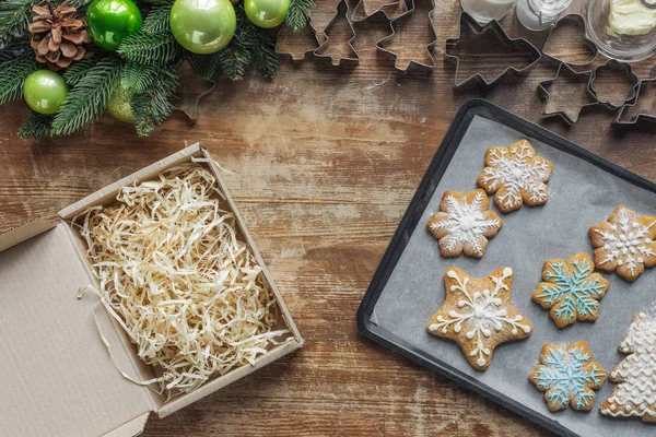 Flat lay com biscoitos de Natal na assadeira, grinalda de Natal, caixa de papelão e cortadores de biscoitos na superfície de madeira — Fotografia de Stock