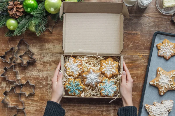 Cropped shot of woman holding box with  homemade christmas cookies on wooden tabletop with decorative christmas wreath — Stock Photo