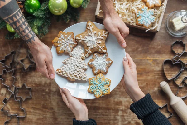 Partial view of man and woman holding plate with homemade cookies on wooden tabletop with decorative christmas wreath — Stock Photo