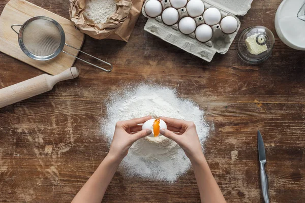 Vista dall'alto di mani femminili che rompono l'uovo in farina su tavolo di legno — Foto stock