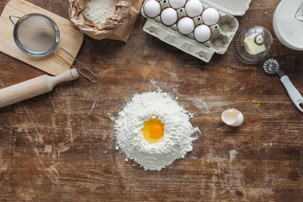 Top view of white flour pile with egg and baking ingredients on wooden table — Stock Photo
