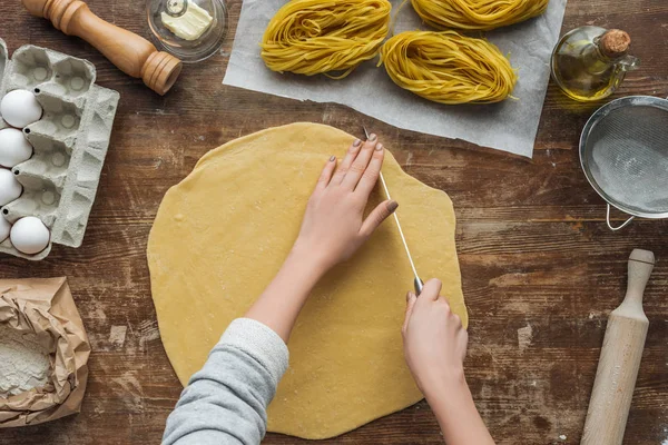 Вид зверху на жіночі руки, що ріжуть тісто для макаронних виробів на дерев'яному столі — стокове фото