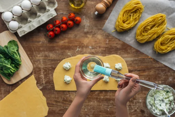 Top view of female hands with bowl and basting brush cooking ravioli above wooden table — Stock Photo