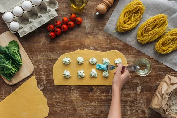 Vue de dessus des mains féminines huilant la pâte pour les raviolis avec brosse à badigeonner à la table en bois — Photo de stock