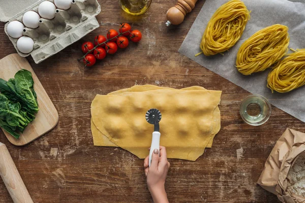 Ansicht von weiblichen Händen, die ein Gebäckrad halten, um Ravioli am Holztisch zu schneiden — Stockfoto