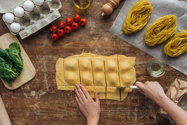 Vista superior de mãos femininas cortando ravioli com roda de pastelaria na mesa de madeira — Fotografia de Stock