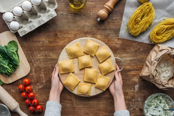 Vista superior das mãos femininas segurando tábua de corte com ravioli na mesa de madeira — Fotografia de Stock