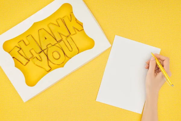 Cropped person holding pen above notebook and square-shaped cake in box with thank you lettering isolated on yellow background — Stock Photo