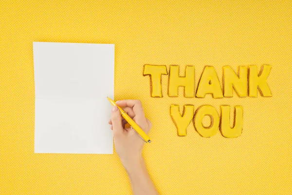 Cropped person holding pen above blank sheet and thank you lettering in cookies isolated on yellow background — Stock Photo