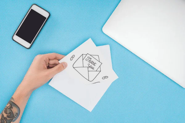 Pessoa cortada segurando cartão branco com obrigado lettering saindo do envelope desenhado à mão e smartphone isolado no fundo azul — Fotografia de Stock