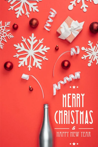 Vista dall'alto del presente, bottiglia di champagne, giocattoli natalizi rossi, nastri bianchi e fiocchi di neve decorativi disposti isolati su rosso con scritte 