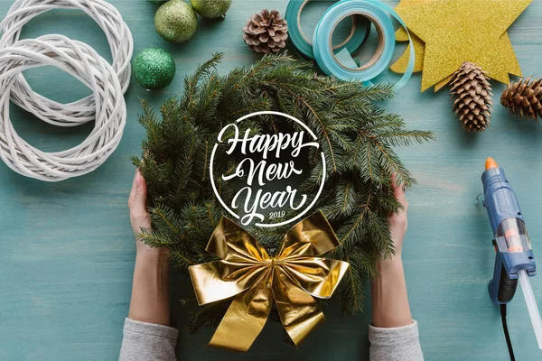 Cropped shot of woman holding handmade pine tree wreath with golden bow and decorations on blue wooden tabletop with 