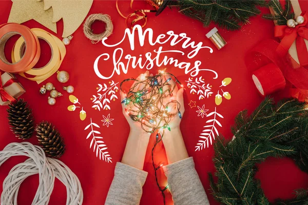 Cropped shot of woman holding christmas lights in hands with pine tree branches and decorations for handmade wreath on red backdrop with 