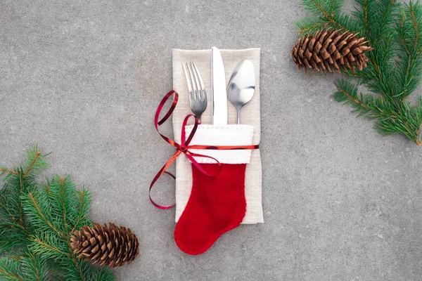 Vista dall'alto della forchetta, cucchiaio con coltello in calza di Babbo Natale avvolto da nastro rosso su tavola con pigne e rami — Foto stock