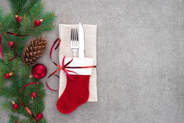 View from above of fork and knife in santa sock wrapped by red ribbon on table with pine cone and branch decorated by christmas balls — Stock Photo