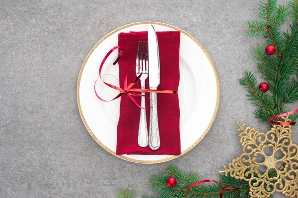 Vue du dessus de l'assiette servie avec fourchette et couteau enveloppé d'un ruban rouge sur la surface avec des branches persistantes décorées par des boules de Noël et une étoile dorée — Photo de stock