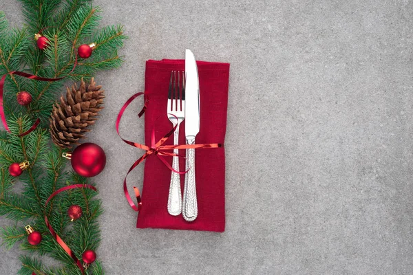Vista superior de tenedor y cuchillo envuelto por cinta roja en la mesa con cono de pino y rama decorada con bolas de Navidad — Stock Photo