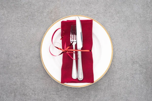 Deitado plano com placa servida com garfo e faca envolto por fita festiva vermelha na superfície cinza — Fotografia de Stock