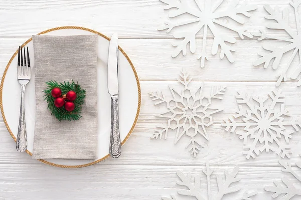 Elevated view of plate with fork, knife and evergreen branch with christmas balls surrounded by decorated snowflakes — Stock Photo