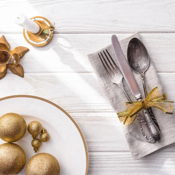Vista elevada de la placa con bolas de navidad doradas, vela y tenedor, cuchara, cuchillo envuelto por cinta en mesa de madera - foto de stock