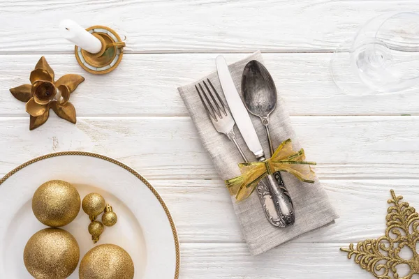 Vista desde arriba del plato con bolas de navidad doradas, vela y tenedor, cuchara, cuchillo envuelto en cinta sobre mesa de madera - foto de stock