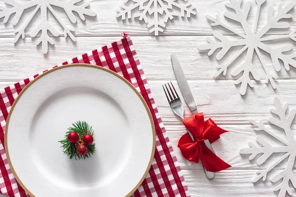 Flat lay with evergreen branch, christmas balls on plate near fork and knife wrapped by red festive ribbon surrounded by decorated snowflakes — Stock Photo