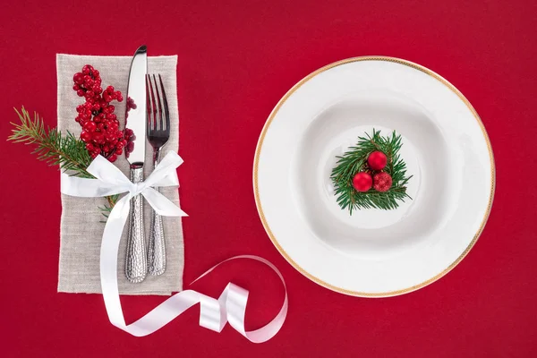 Blick von oben auf Gabel, Messer, rote Beeren mit Band umwickelt und Teller mit immergrünem Baumzweig mit Weihnachtskugeln isoliert auf rot — Stockfoto