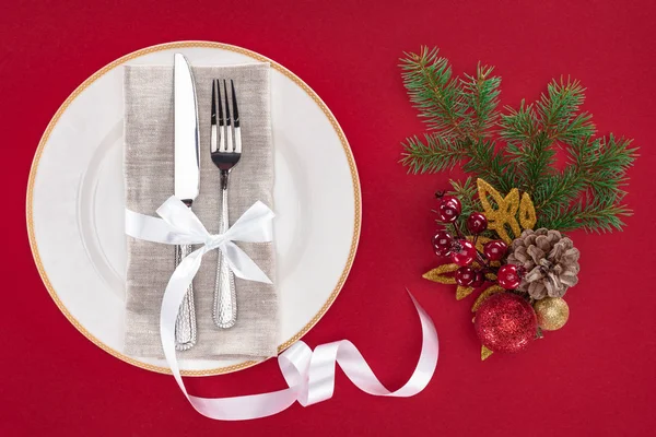 Повышенный вид вилки и ножа, завернутые лентой на тарелке возле рождественских украшений, изолированных на красный — стоковое фото